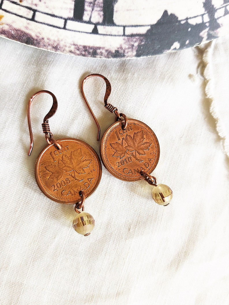 Peut inclure: Une paire de boucles d'oreilles de couleur cuivre avec des pi&egrave;ces de monnaie canadiennes de 1 cent de 2008 et 2010, chacune avec un motif de feuille d'&eacute;rable. Les pi&egrave;ces sont attach&eacute;es &agrave; des perles de verre transparentes.