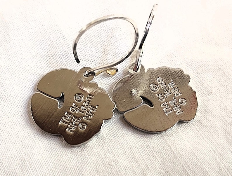 May include: A pair of silver earrings shaped like hockey pucks. The back of the earrings are engraved with the text "TM & &copy; NHL Team NHL"