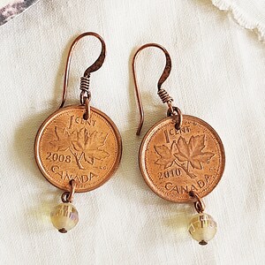 Peut inclure: Une paire de boucles d'oreilles en cuivre avec des pi&egrave;ces de monnaie canadiennes de 1 cent de 2008 et 2010, chacune avec un motif de feuille d'&eacute;rable et une perle de cristal transparente.