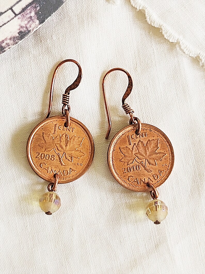 Peut inclure: Une paire de boucles d'oreilles en cuivre avec des pi&egrave;ces de monnaie canadiennes de 1 cent de 2008 et 2010, chacune avec un motif de feuille d'&eacute;rable et une perle de cristal transparente.