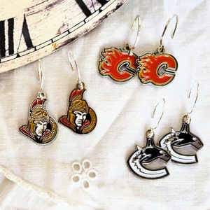 May include: Set of six hockey team themed earrings. Two pairs feature red and white logos of the Calgary Flames, one pair features a black and white logo of the Vancouver Canucks, and two pairs feature gold and black logos of the Ottawa Senators.