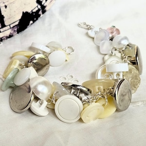 May include: A unique bracelet made from a collection of buttons in white, silver, and pale yellow hues. The buttons are linked together with silver-coloured chains and clasps, creating a distinctive piece of jewellery.
