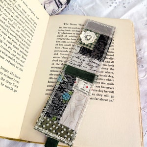 May include: A handmade fabric bookmark with a green, white and black patchwork design. The bookmark has a green ribbon attached to the bottom. The text "The Stone World" is visible on the page behind the bookmark.