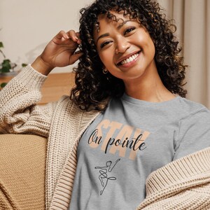 May include: A gray t-shirt with the words "Stay On Pointe" in peach and black script, along with a line drawing of a ballerina. The person is wearing a beige cardigan.