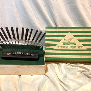 May include: Vintage cocktail fork set with a wooden base and dark brown handles. The forks are arranged in a fan shape within a box with a green interior. The box is next to a green and white striped box that reads "COCKTAIL FORK SET."