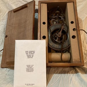 May include: A vintage clock mechanism inside a wooden box, with the lid open. The clock face has Roman numerals. A paper with text is in front of the box. The text on the paper reads "VERY IMPORTANT! KEEP WEDGE AND TUBE TO REPACK Kronos for STORING OR SHIPPING."