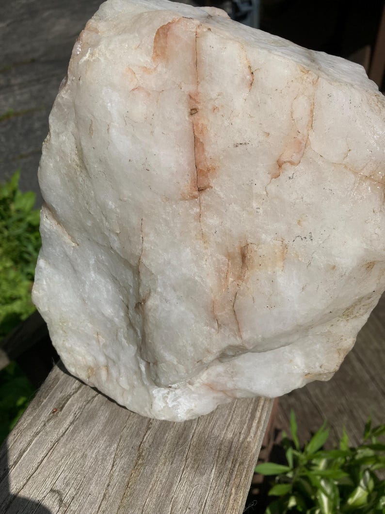 May include: A large, irregularly shaped quartz rock. The rock has a creamy white base colour with reddish-brown veining. It has a rough, textured surface and is displayed outdoors.