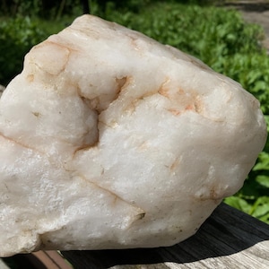 May include: A large, irregularly shaped quartz crystal. The stone has a milky white base with subtle peach-coloured veins. It is displayed on a weathered wooden surface, with green foliage in the background.