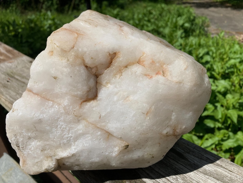 May include: A large, irregularly shaped quartz crystal. The stone has a milky white base with subtle peach-coloured veins. It is displayed on a weathered wooden surface, with green foliage in the background.