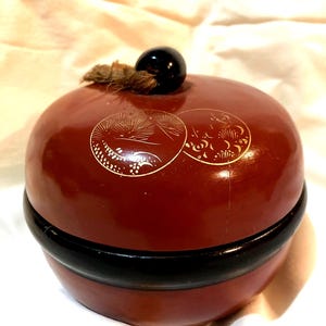May include: A round, lacquered box in a deep red hue, featuring a black base and a glossy black knob on top. The lid is adorned with two circular, gold-colored decorative designs. A small, natural-colored rope detail is attached to the top.