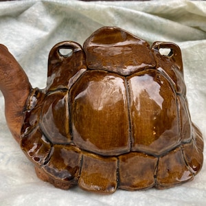 May include: A ceramic turtle-shaped container with a glossy brown finish. The turtle has a detailed shell and a head with an open mouth. The container has two small handles on the top of the shell.