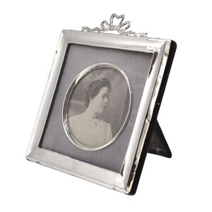 May include: Silver picture frame with a decorative bow at the top. The frame holds a black and white photo of a woman.