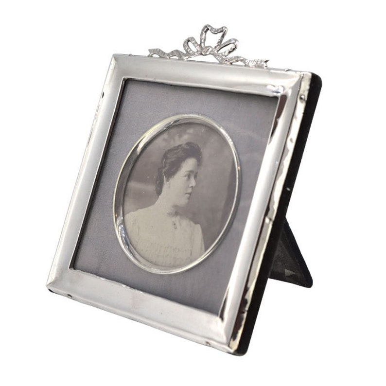 May include: Silver picture frame with a decorative bow at the top. The frame holds a black and white photo of a woman.