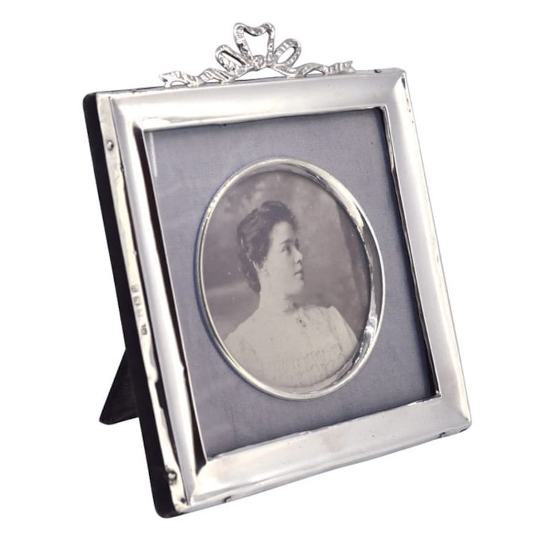 May include: A silver picture frame with a decorative bow at the top. The frame is standing on a black stand and holds a black and white photo of a woman.