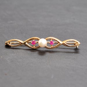 Puede incluir: Broche dorado con un diseño alargado y simétrico. Presenta una perla blanca central, flanqueada por dos pequeñas piedras rojas redondas. El broche tiene un intrincado patrón calado.