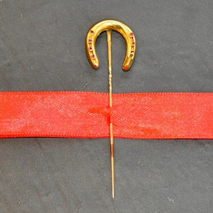 May include: A gold horseshoe pin with red gemstones. The pin is on a red ribbon.