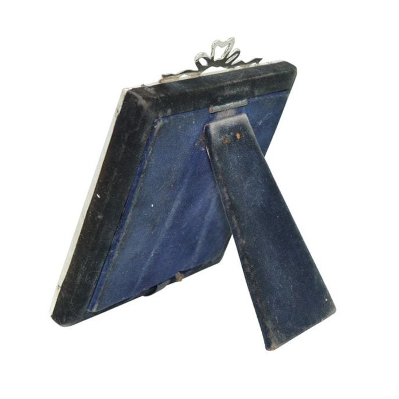 May include: A silver-toned picture frame with a decorative design on the top. The frame is standing on a blue velvet stand.