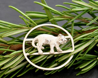 Könnte beinhalten: Eine silberne Brosche mit einer stilisierten Katze, die in einem Kreis mit einem dekorativen Design geht. Die Brosche liegt auf einem Bett aus grünem Rosmarin.