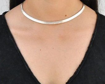 Può includere: Una collana a catena d'argento con un design semplice ed elegante. La catena è sottile e piatta e si appoggia vicino al collo.
