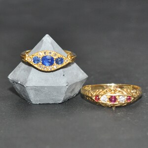 Op de afbeelding: Twee gouden ringen met edelstenen. De eerste ring heeft drie blauwe saffieren en kleine diamanten die in een gouden band zijn gezet. De tweede ring heeft drie rode robijnen en kleine diamanten die in een gouden band zijn gezet.