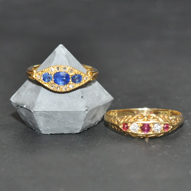 Op de afbeelding: Twee gouden ringen met edelstenen. De eerste ring heeft drie blauwe saffieren en kleine diamanten die in een gouden band zijn gezet. De tweede ring heeft drie rode robijnen en kleine diamanten die in een gouden band zijn gezet.