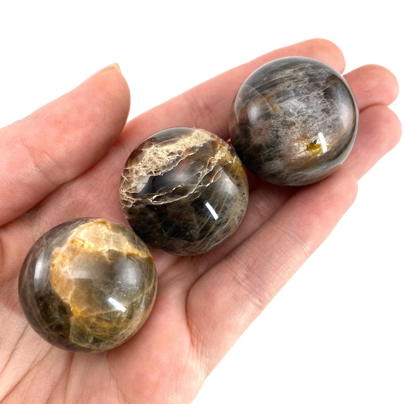 May include: Three polished black and gold gemstone spheres, also known as sunstone spheres, are displayed in a hand.