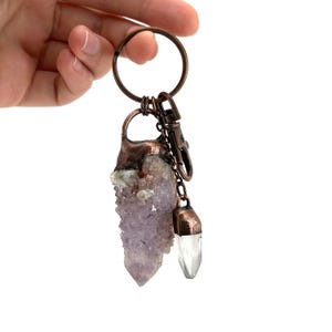 May include: A copper-toned keychain featuring a large, light purple amethyst crystal and a smaller, clear quartz crystal pendant. The crystals are attached with a chain and clasp to a round key ring. The crystals have a natural, textured appearance.