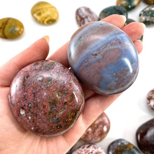 May include: A hand holds two smooth, oval-shaped stones. One is a deep red with green and brown inclusions, the other has a blue and maroon swirl pattern. Other stones are visible in the background. The stones are polished and have a glossy finish.