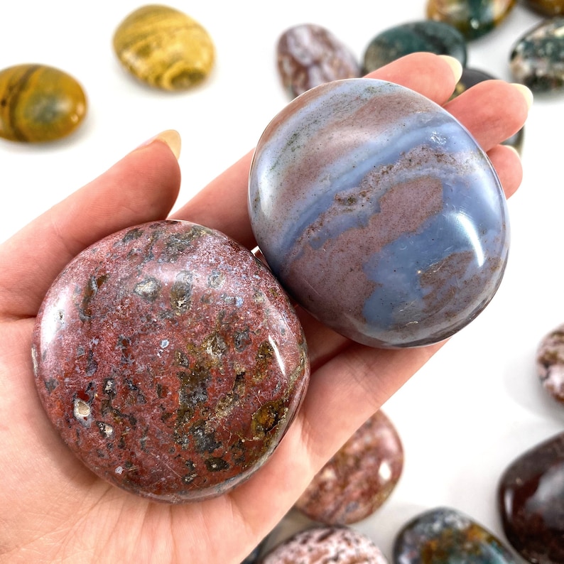 May include: A hand holds two smooth, oval-shaped stones. One is a deep red with green and brown inclusions, the other has a blue and maroon swirl pattern. Other stones are visible in the background. The stones are polished and have a glossy finish.