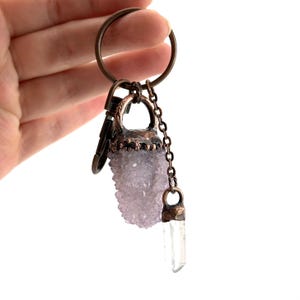 May include: A bronze-toned keychain featuring a cluster of light purple crystals and a clear quartz crystal pendant. The crystals are attached with a chain and a clasp. The keychain is held against a white background.