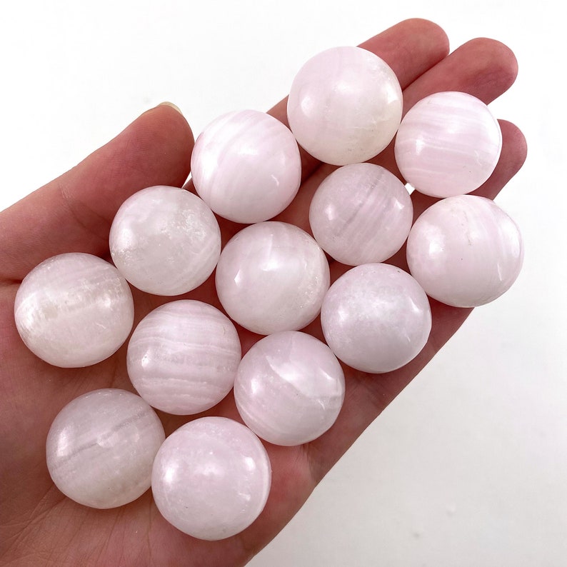 May include: A collection of 12 smooth, white selenite spheres with a subtle pink hue. The spheres are polished and have a striated texture.
