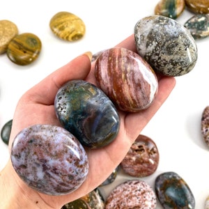 May include: A selection of polished, oval gemstones in shades of brown, white, green, and purple. The stones are held in a hand, with more scattered around. The gemstones display unique patterns and textures, ideal for collectors or decorative use.
