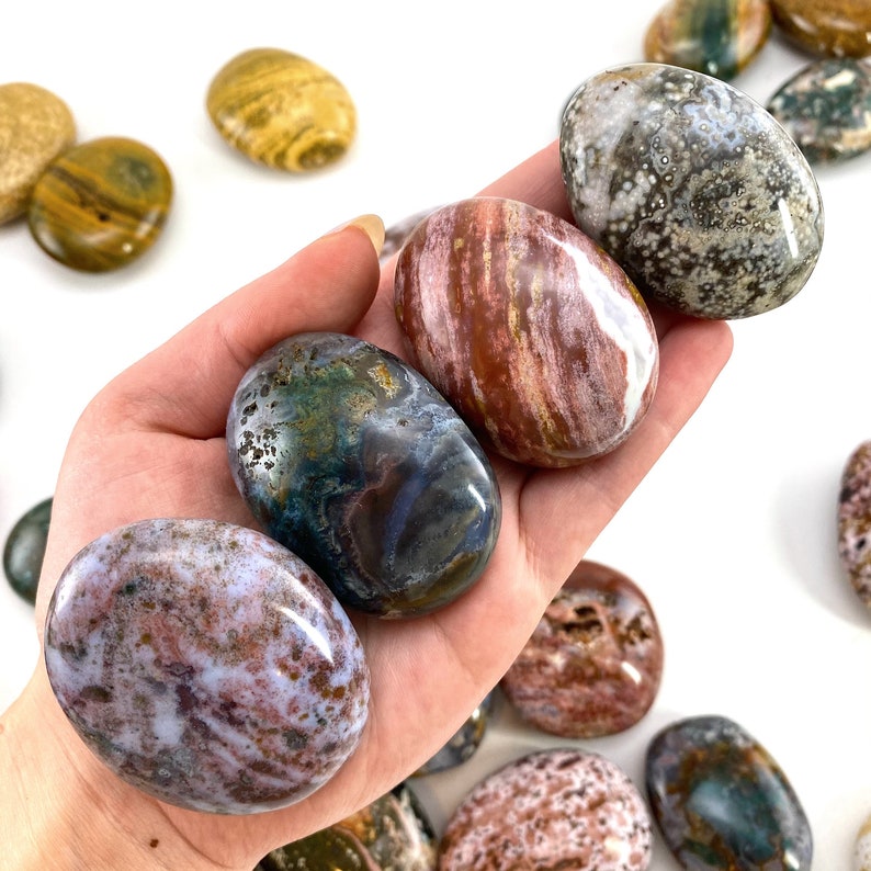 May include: A selection of polished, oval gemstones in shades of brown, white, green, and purple. The stones are held in a hand, with more scattered around. The gemstones display unique patterns and textures, ideal for collectors or decorative use.