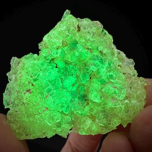 Puede incluir: Primer plano de un espécimen mineral verde brillante sobre un fondo negro. La estructura cristalina tiene una apariencia burbujeante y agrupada, con áreas de intensa luminosidad. La forma general es irregular, mostrando formaciones naturales. El color es verde brillante, casi neón.