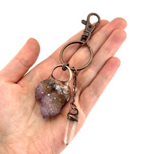 May include: A copper-colored keychain featuring a raw amethyst crystal and a clear quartz crystal pendant. The amethyst is a light purple color, and the quartz is clear. The keychain has a lobster clasp and a chain connecting the crystals.