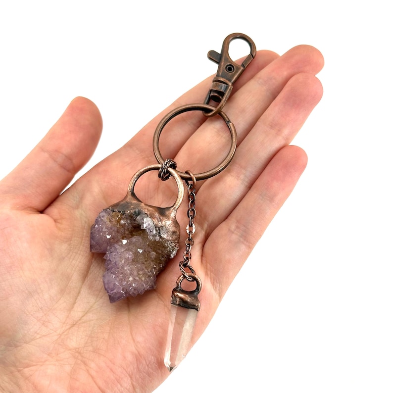 May include: A copper-colored keychain featuring a raw amethyst crystal and a clear quartz crystal pendant. The amethyst is a light purple color, and the quartz is clear. The keychain has a lobster clasp and a chain connecting the crystals.