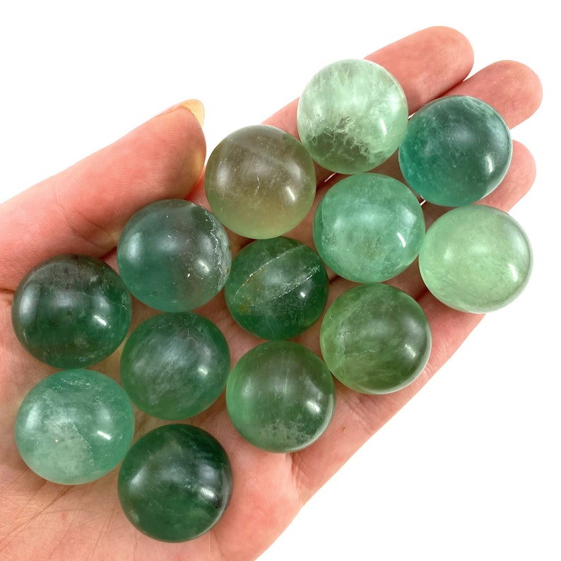 May include: A collection of 13 green fluorite spheres, each with a slightly different shade of green. The spheres are smooth and polished, and they are arranged in a loose pile.