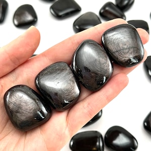 May include: A collection of black obsidian tumbled stones with a silver sheen. The stones are smooth and polished, and they have a variety of shapes and sizes.