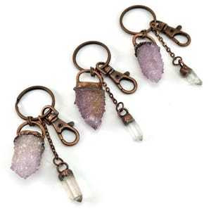 May include: Three keychains with amethyst crystal clusters and clear quartz crystal pendants. The keychains have bronze-colored metal rings, clasps, and chain accents. The amethyst crystals are a light purple color.
