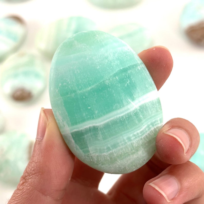 Op de afbeelding: Een gladde, ovale groene en witte celestiet palmstone. De steen heeft een lichtgroene kleur met witte strepen.