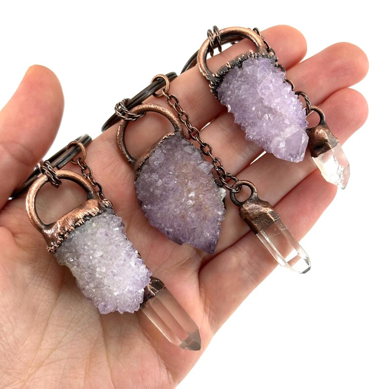 May include: Three crystal pendants with copper-colored metal accents. Each pendant features a cluster of light purple crystals and a clear quartz crystal point. The pendants are connected by a black cord.