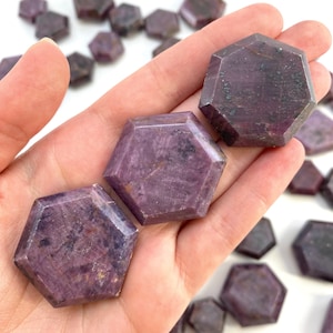 May include: Three purple hexagonal shaped gemstones, also known as purple sapphires, are held in a hand. The gemstones are faceted and have a smooth, polished finish.