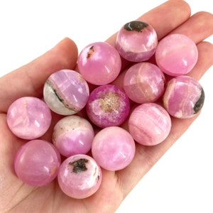 May include: A collection of pink and white tumbled spheres of pink calcite, a variety of calcite that is known for its pink color and white veins. The spheres are smooth and polished, and they are arranged in a hand.