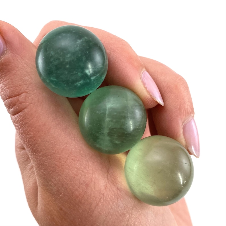 May include: Three green gemstone spheres, each with a different shade of green, are held in a hand. The spheres are smooth and polished.