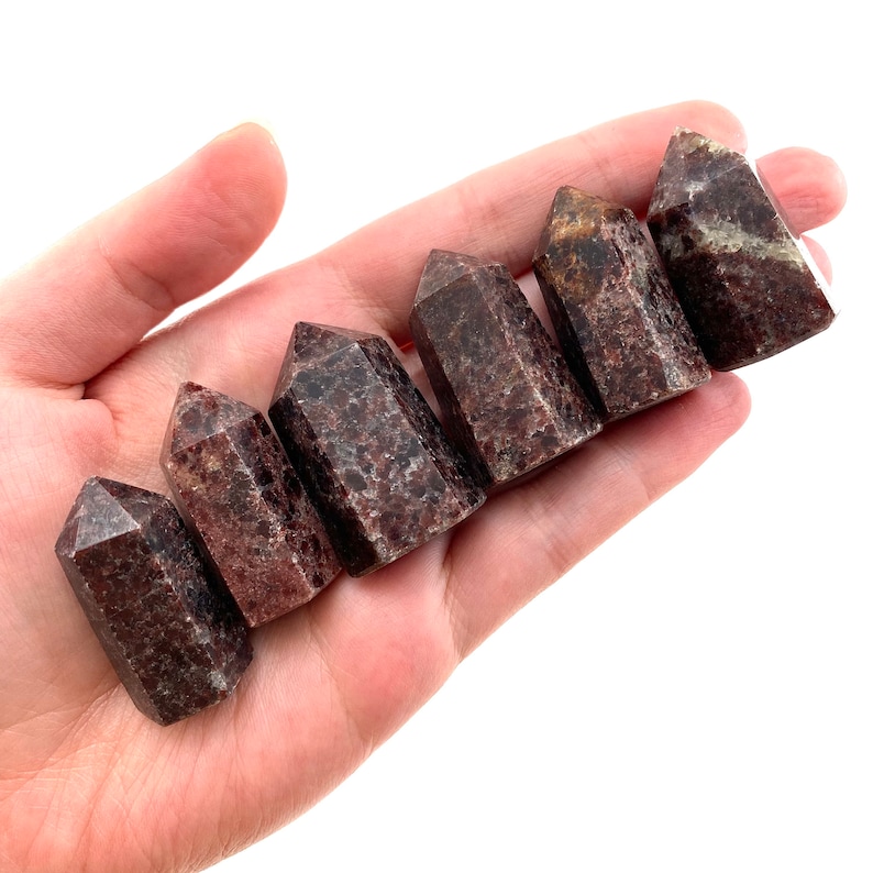 May include: Seven red and brown gemstone points, also known as red jasper points, are arranged in a row on a hand. The points are all different sizes and shapes, but they are all polished and have a smooth, shiny finish.