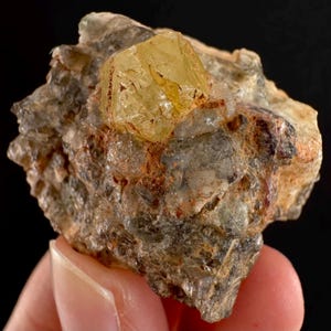 Może przedstawiać: Surowy, naturalny okaz minerału z wyraźnym, fasetowanym, żółto-pomarańczowym kryształem. Kryształ osadzony jest w matrycy szarej, brązowej i czerwonobrązowej skały. Ogólna tekstura jest szorstka i nierówna, z widocznymi strukturami krystalicznymi.