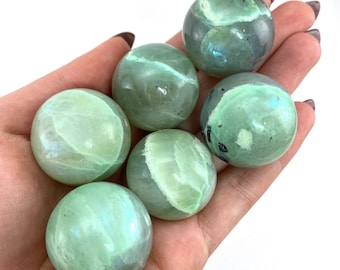 May include: Six green and white gemstone spheres, also known as orbs, are displayed in a hand. The spheres are smooth and polished.