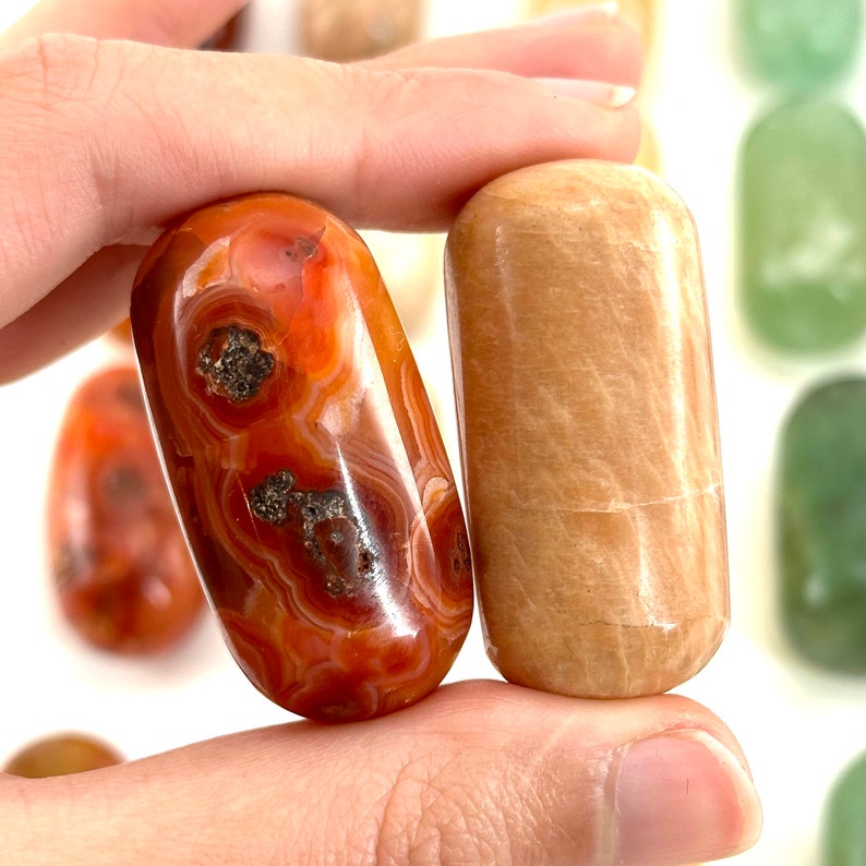 May include: Two polished oval-shaped stones, one is a carnelian with orange and red banding and the other is a light brown selenite.