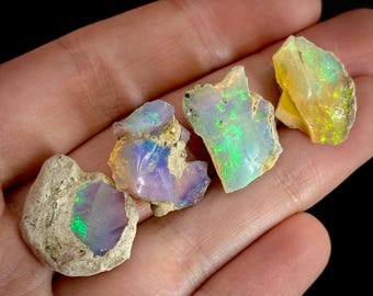 May include: Four raw opal gemstones with vibrant colors of green, yellow, and purple. The opals are rough-cut, showcasing their natural, unpolished textures. The gemstones are held in a hand against a dark background.