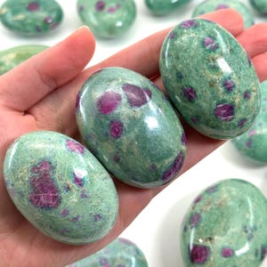 May include: Several oval-shaped stones with a green base color and vibrant red inclusions. The stones are smooth and polished, with a natural, organic appearance. The stones are held in a hand, and are likely gemstones or decorative items.